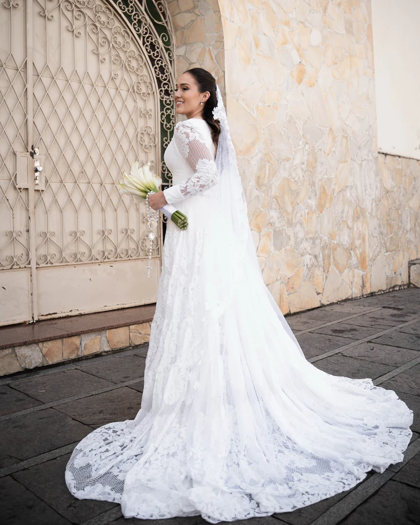 Noiva de corpo inteiro em vestido com cauda rendada em frente ao portal da igreja